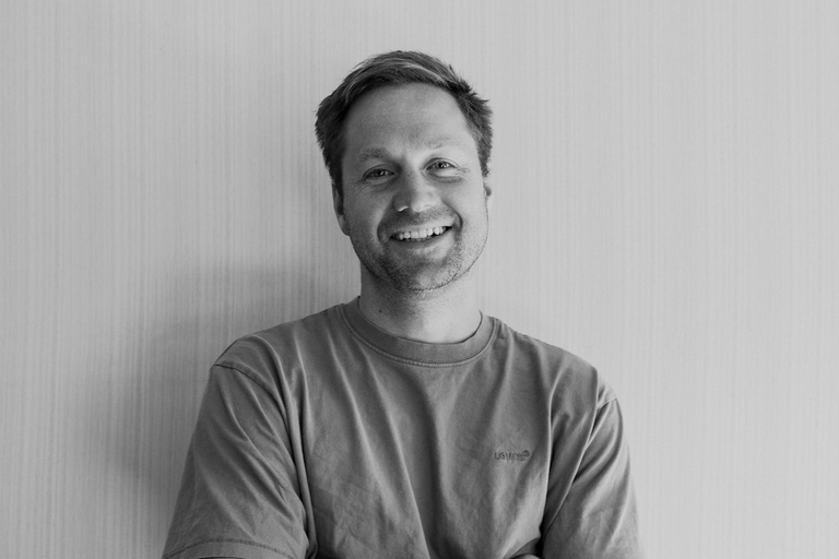 Black and white headshot of Harry Moore smiling in a casual t-shirt against a plain backdrop, ideal for a professional portrait or bio page.